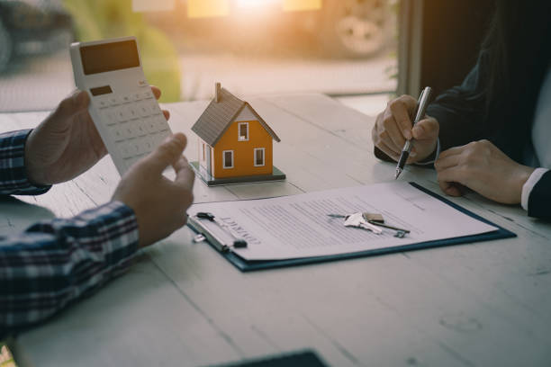 Two people working on a real estate contract with a small house model, calculator, and keys on the table.