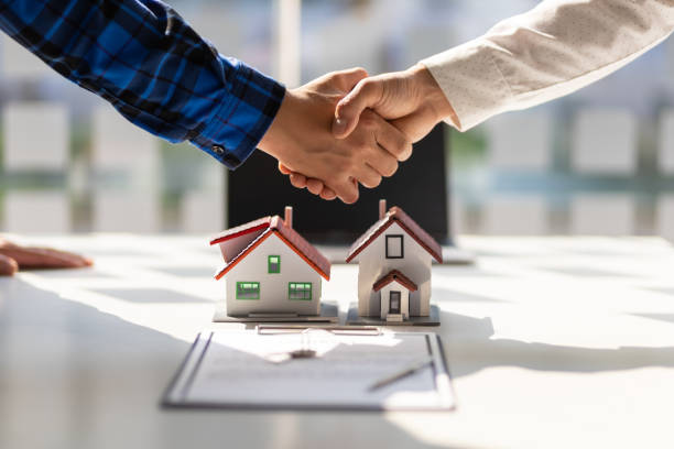 Two people shaking hands over a table with small house models and a contract.