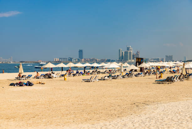 Sunbathers on a sandy beach in Dubai with skyscrapers visible in the background under a clear blue sky.