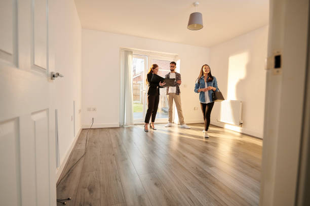 Three people touring an empty, sunlit room with wooden flooring in a house.