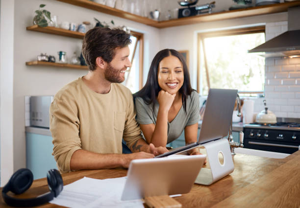 A smiling couple works together on laptops at a kitchen table.